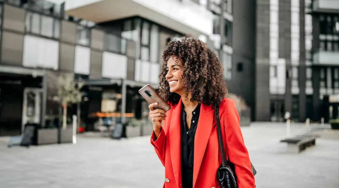 A smiling woman in a red blazer holding a smartphone in an urban setting, embodying confidence and modern lifestyle.