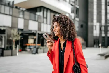 A smiling woman in a red blazer holding a smartphone in an urban setting, embodying confidence and modern lifestyle.