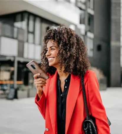 A color image of a smiling woman standing outdoors in an urban setting. She has curly hair and is wearing a bright red blazer over a black top, holding a smartphone in her hand. The background shows modern buildings, slightly blurred, giving the image a professional and contemporary feel. The woman’s expression is joyful and confident, suggesting a positive and upbeat mood. Her attire and demeanor convey a sense of professionalism and style.






