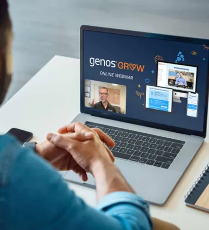 A color image of a person watching a webinar on their laptop. The laptop screen displays the title "Genos Grow Online Webinar" with an ongoing presentation. A presenter is visible in a small video frame, and there are additional visuals showing content from the webinar, such as slides and software interfaces. The viewer’s hands are clasped in front of the laptop, indicating attention and engagement. A smartphone and a notebook with a pen are placed nearby on the table, suggesting a professional or learning environment.







