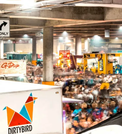 A vibrant color image of a bustling night market under a covered parking area. The scene is filled with a large crowd of people moving around food trucks and vendor stalls. Brightly lit food trucks display colorful signs, such as "Dirtybird" and "G-Dup," offering a variety of food options. The atmosphere is energetic, with blurred motion capturing the lively activity and movement of people. Overhead lights illuminate the space, creating a festive and welcoming environment. The image conveys the excitement and dynamic nature of the night market experience.






