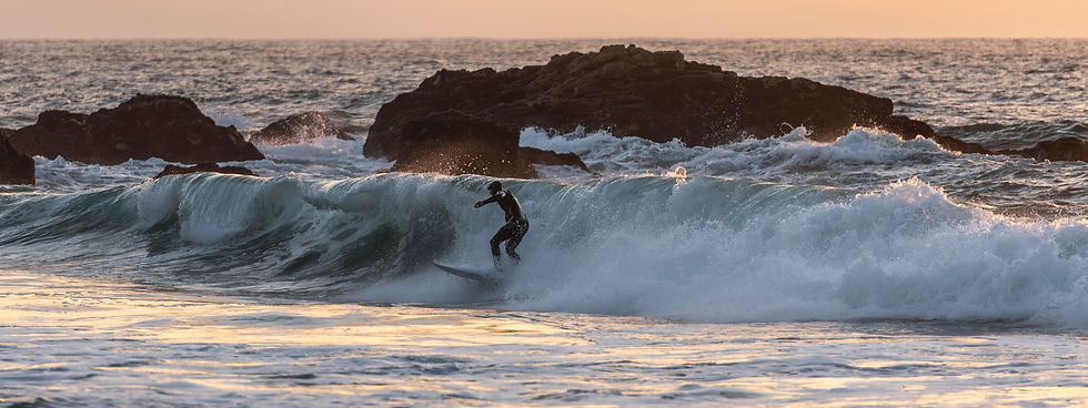 Surfer riding a breaking wave near dark coastal rocks at sunset, light fading as motion continues.
