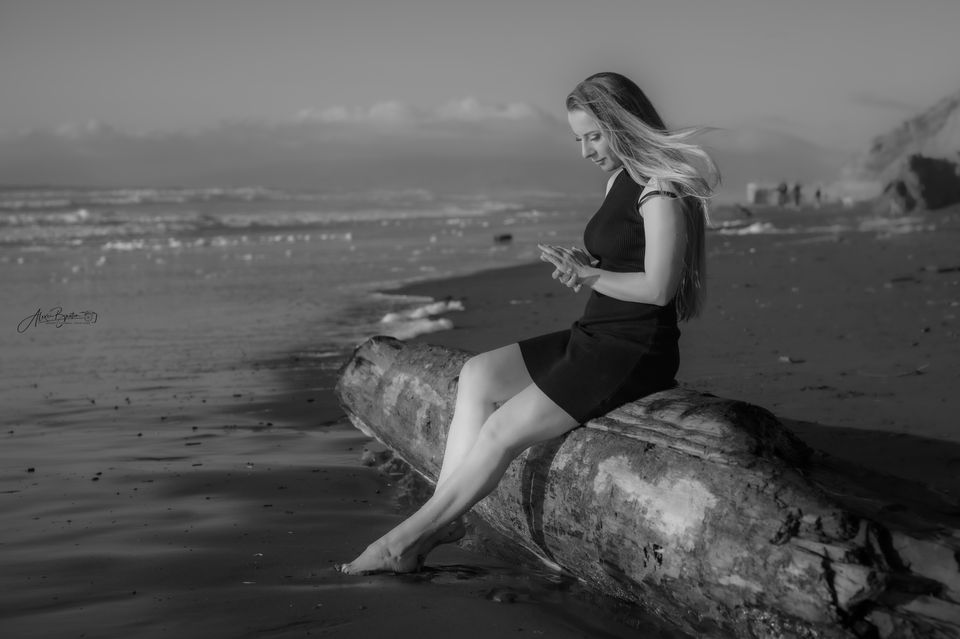 Black and white portrait of a lady sitting on a log by the ocean.