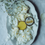 Thumbnail: yellow arnica relief balm in a gold tin on flowers
