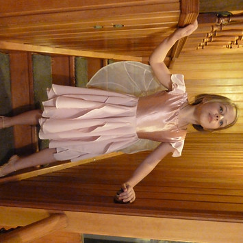 A girl stands on wood stairs in a pink dress with fairy wings.