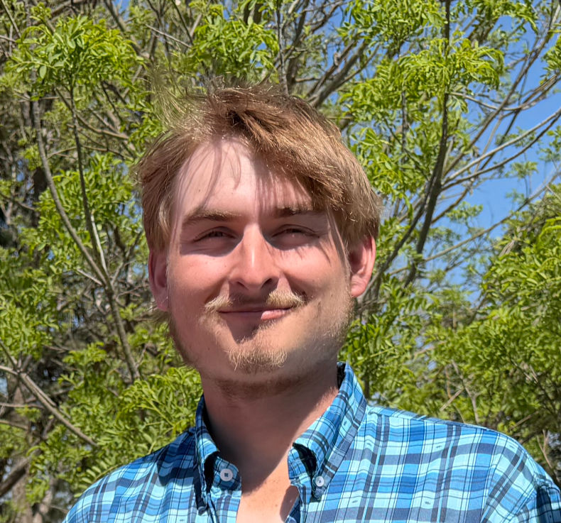Smiling man in blue plaid shirt outdoors with green trees