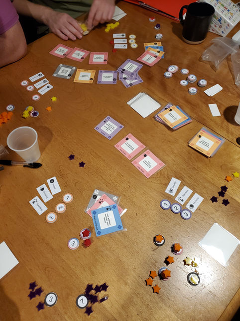 Four-player game of cutting flowers on a wooden table. Lots of cards in 5 different colors as well as tokens spread across the table.