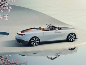 Convertible Rolls Royce Drop tail car parked on a light concrete surface beside a reflective water body. Pink blossoming tree nearby. Calm and serene mood.