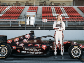 Female race car driver standing confidently in front of a Charlotte Tilbury–sponsored Formula 1 car decorated with bold lipstick graphics, on a professional racetrack.