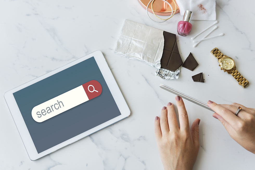 Hands filing nails on a marble table with a tablet showing "search," chocolate, nail polish, cotton swabs, and a gold watch nearby.