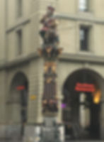 A photo of a statue.  The statue has a larger man at the top, and he is depicted as eating babies.  There are babies crawling around him, and he has one in his hand near his mouth, and his mouth is open.  In the background, a sign says "Kornhaus Restaurant".  There are multiple fountains at the bottom of the statue