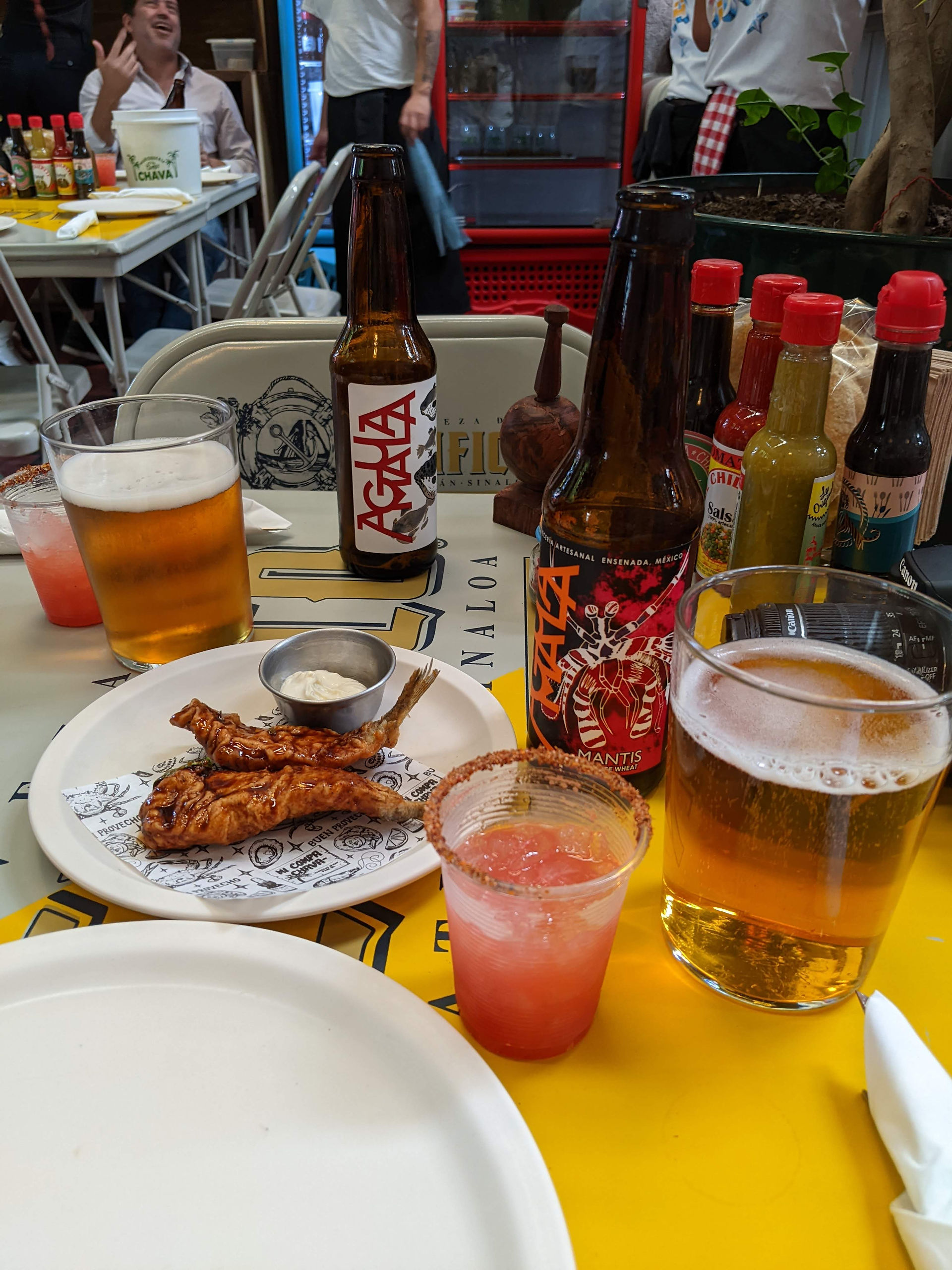 photo of 2 pieces of fried fish on a plate, two beers poured out into glasses, and a red colored drink with seasoning on the tip