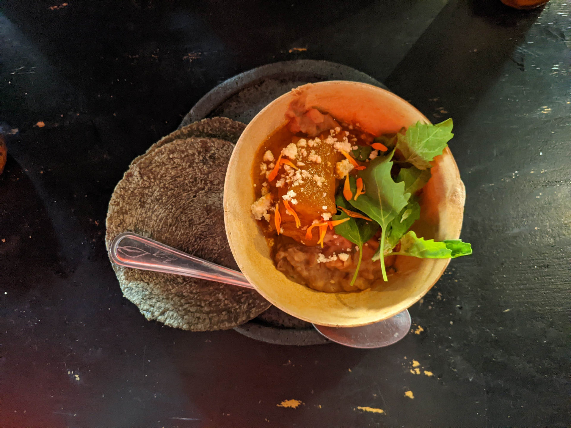 black plate with bowl on top with meat and sauce and green lettuce.  To the left, a blue corn tortilla.  A spoon is to the bottom of the plate.