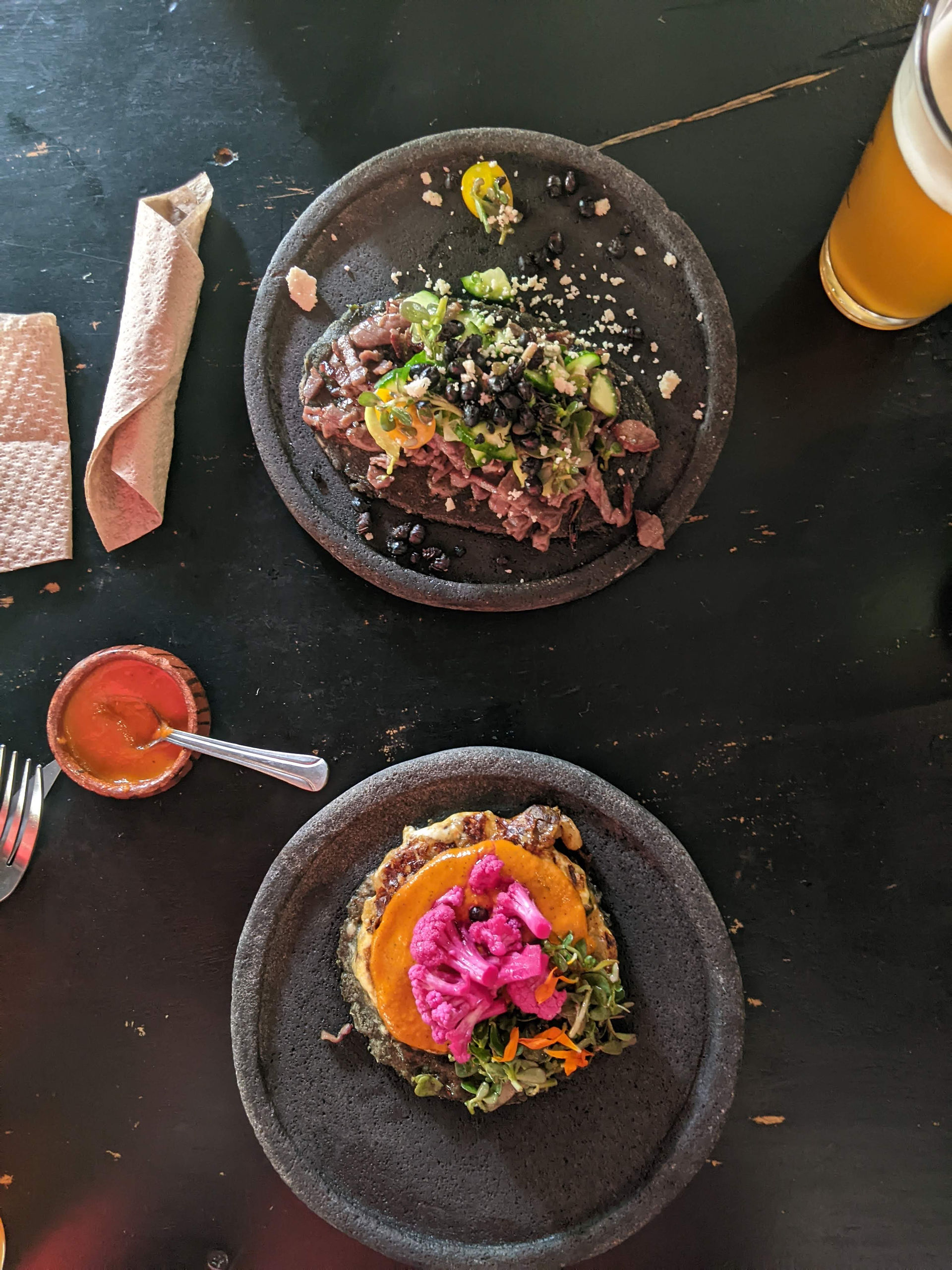 2 black plates both containing different tacos. Taco on bottom plate has red pickled cauliflower and an orange sauce. Top plate has cooked insects on it along with other toppings