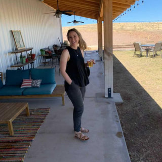 A woman is on a patio, smiling at the camera. She has a cup in her hand, her other hand in her pocket. She is wearing sandals, black pants, and a black tank top.