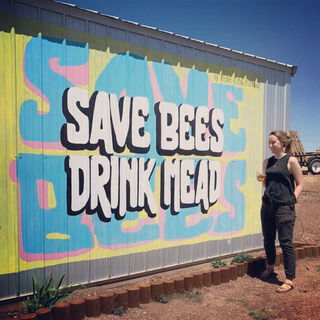 The large colorful mural on the side of a small building reads, "Save bees, Drink Mead". A woman stands next to it with a cup in her hand. She is wearing sandals, black pants, and a black tank top.