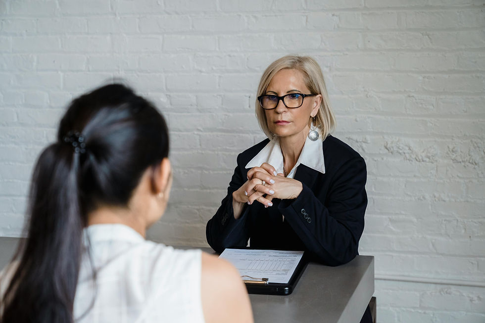 Two women in an interview setting, one facing the camera with folded hands, the other with a ponytail, against a white brick wall.