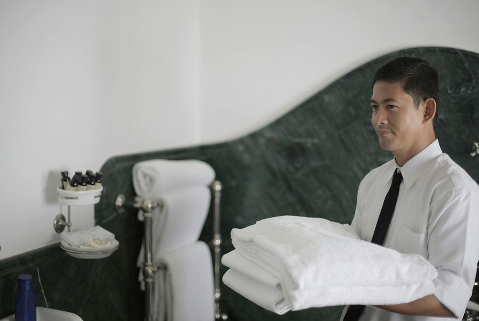 Man holding folded white towels in a white and green tiled bathroom. Soap and toiletries on the left. Calm expression.