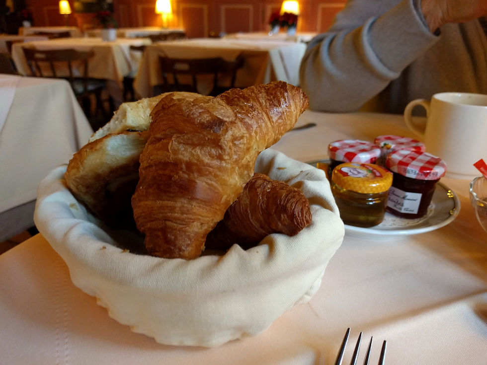 Basket of croissants on a restaurant table with jam jars, a mug, and a fork. Warm, cozy lighting with beige tablecloths in the background.
