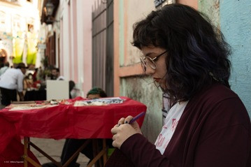 A foto mostra Juliana sentada, encostada em uma parede à direita do enquadramento. Ela olha para baixo em direção às mãos enquanto costura ou borda.