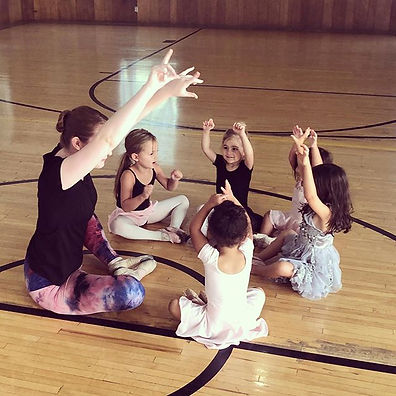 Miss Eden sits with a group of preschool ballet students as they all reach their hands to the sky.