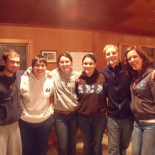Six young people in sweatshirts and jeans smiling with arms around each other in a cabin