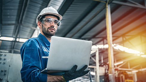 A safety-clad worker in an industrial setting, holding a laptop with illuminated machinery in the background, symbolizing technology integration and sustainable operations.