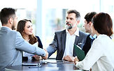 Two young men in business-casual attire shake hands across a table as other young men and women look on approvingly.