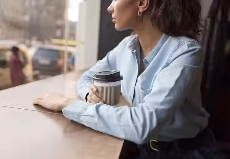 uma mulher a olhar pela janela refletindo, com uma caneca de café
