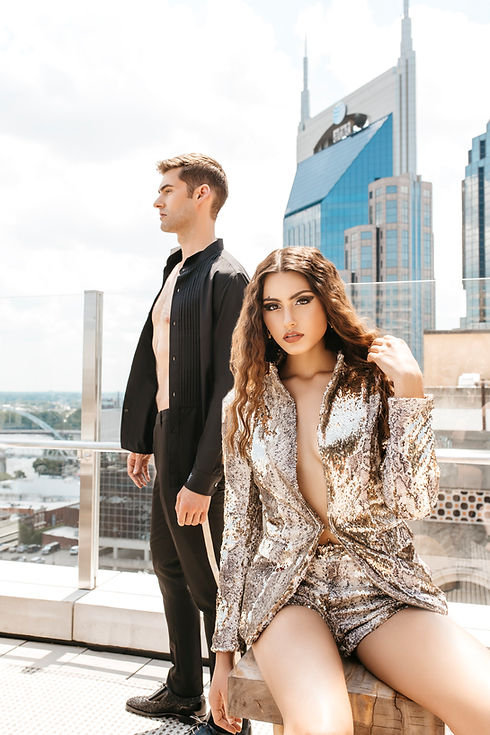 A woman in a silver sequined outfit poses seated while a man in a black suit stands behind her on a rooftop with Nashville skyscrapers in the background. Captured by Weatherly Photography, the image epitomizes chic city lifestyle.
