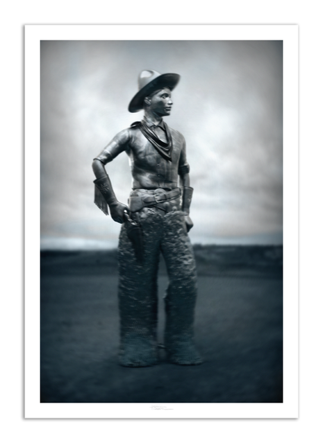 A fine art print by Trent Tate showing a cowboy standing with his gloved hand holding the pistol in its holster on his hip. He is wearing boots and spurs, woolly chaps, a bandana, and his sleeves are rolled up. He stares into the distance intently.