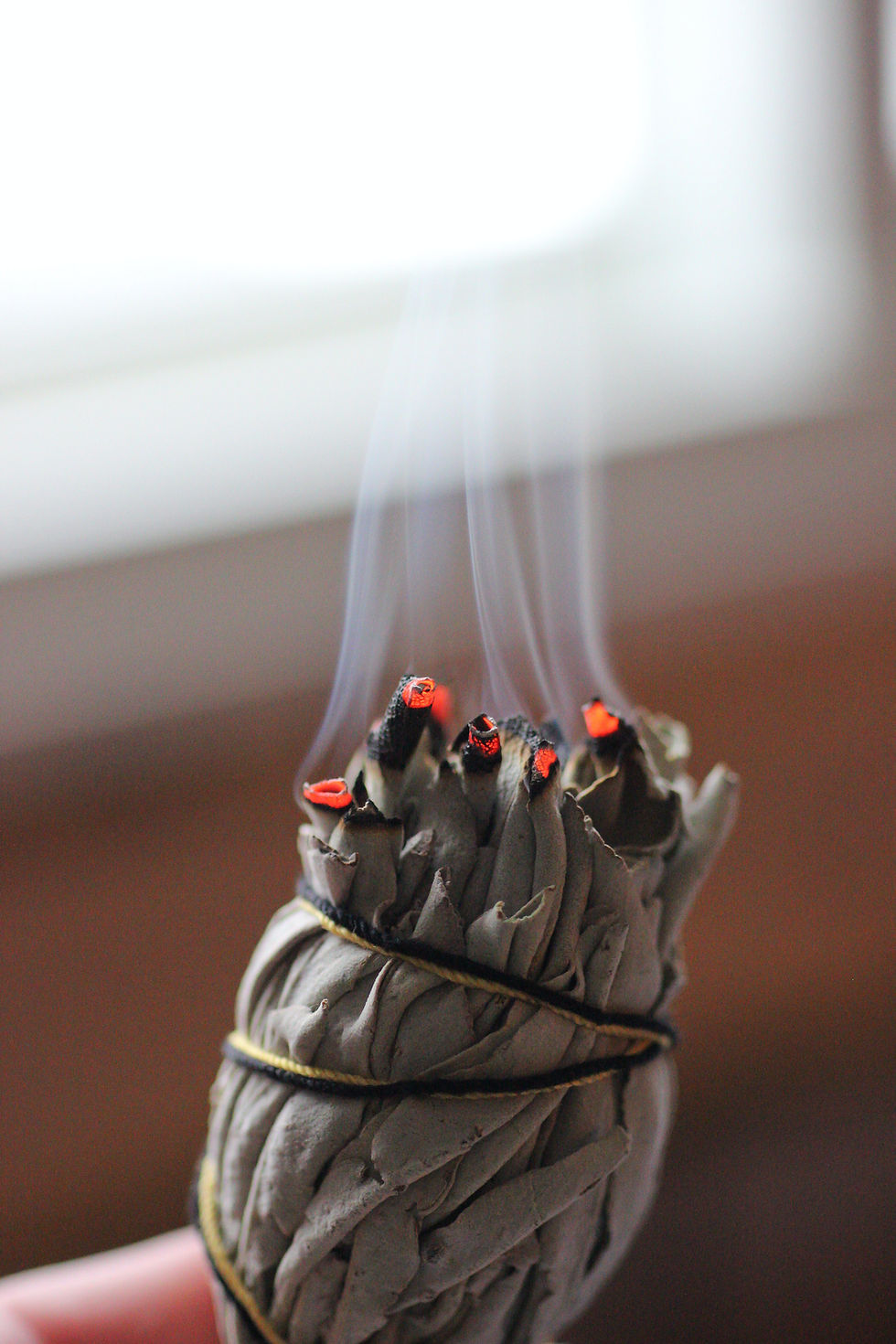A smoking white sage smudge bundle, the holder's finger just in view.
