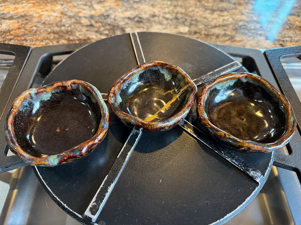 Three matching handmade ceramic sauce bowls arranged side by side on a black stovetop burner grate. The center bowl shows a visible golden kintsugi repair line — the one chosen, always, for its story.