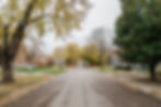 A quiet residential street lined with mature trees in autumn color, modest brick and clapboard houses set back from the road, fallen leaves scattered across the pavement and sidewalks under an overcast sky.