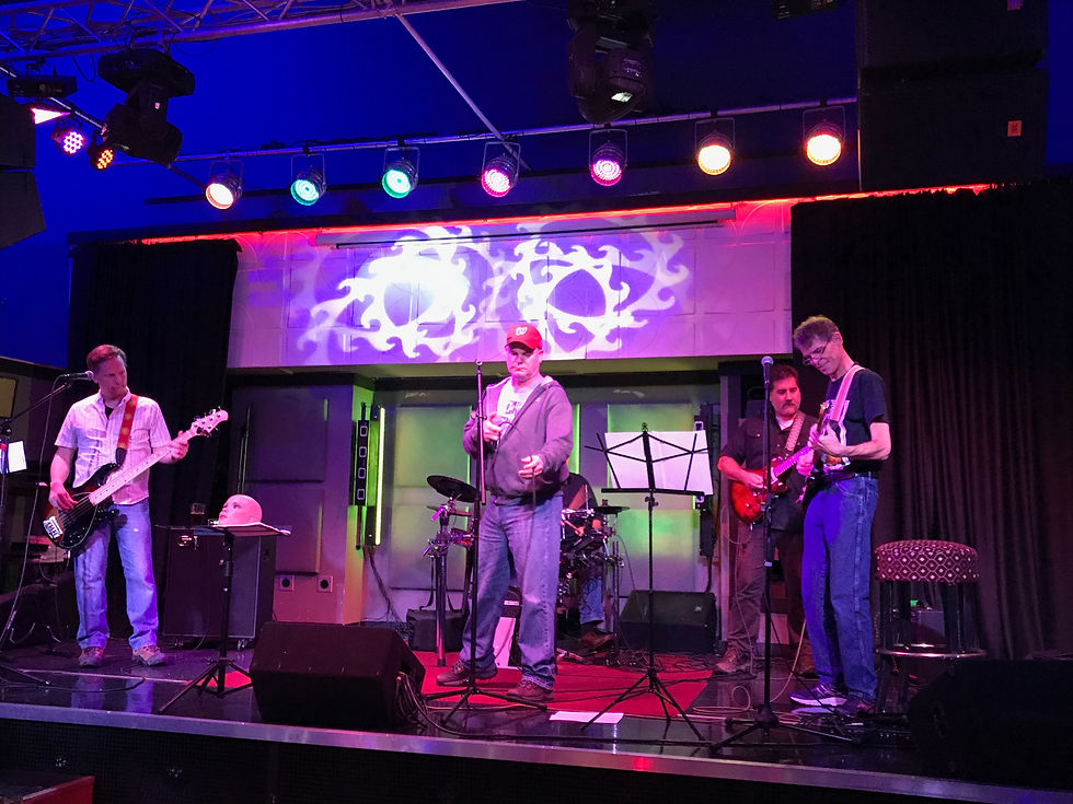 Four musicians perform on a small club stage under colorful lights — the guitarist on the far right in a black t-shirt and jeans, focused intently on playing, is Chip.