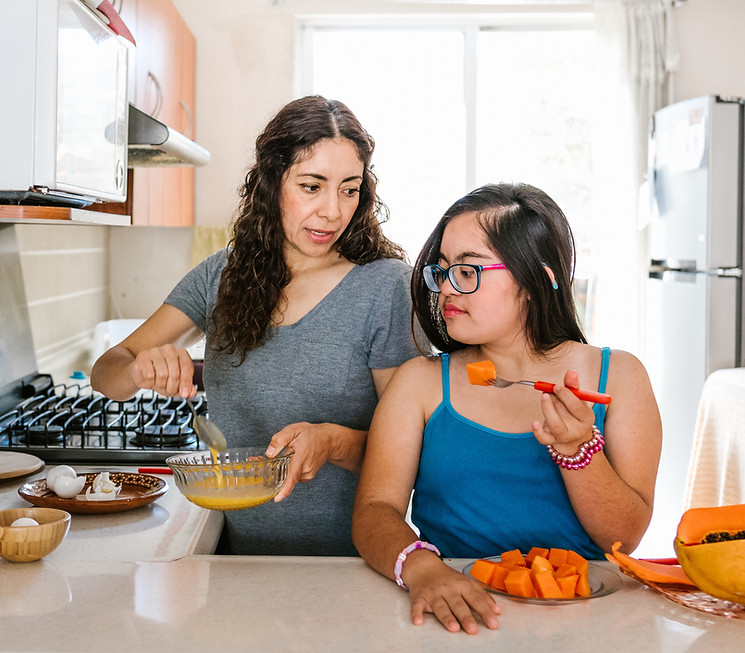 Mexican teen girl with down syndrome eating fruit and her mother cooking at home, in disab