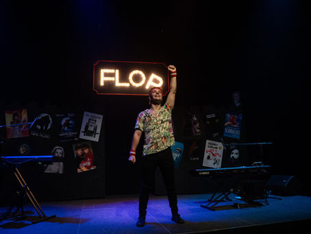 Performer on a dark stage raises a glowing FLOP sign, with musical posters and keyboards behind him.