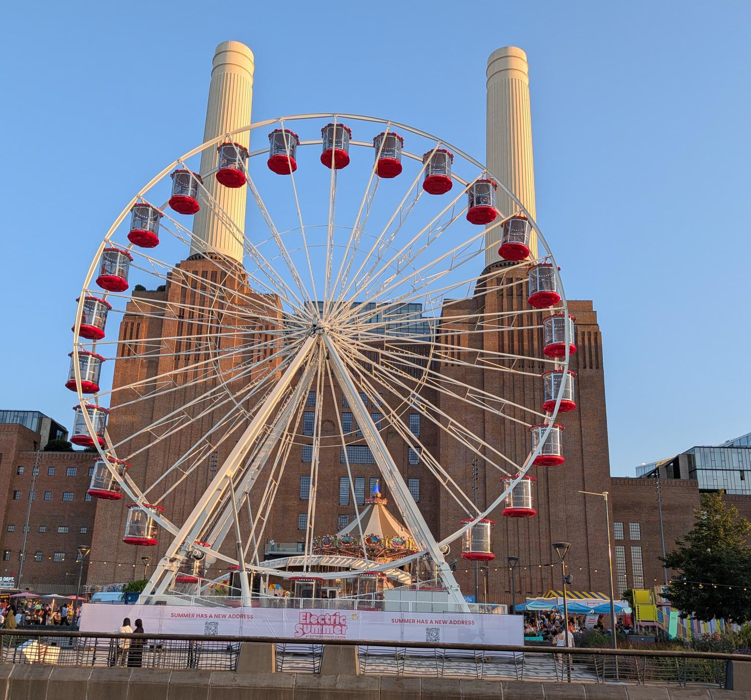 Ferris wheel with red gondolas in front of a brick power station, banner reading Electric Summer under a clear blue sky.