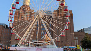 Ferris wheel with red gondolas in front of a brick power station, banner reading Electric Summer under a clear blue sky.