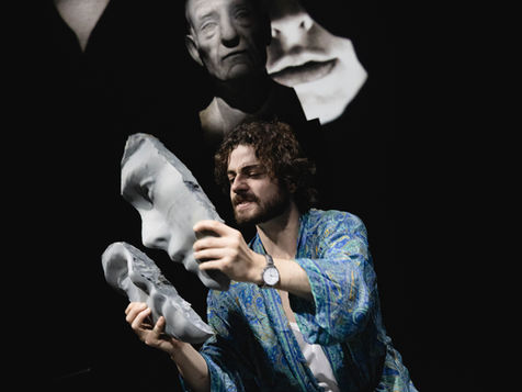 A man in a dressing gown holds pieces of a broke sculpture's face.