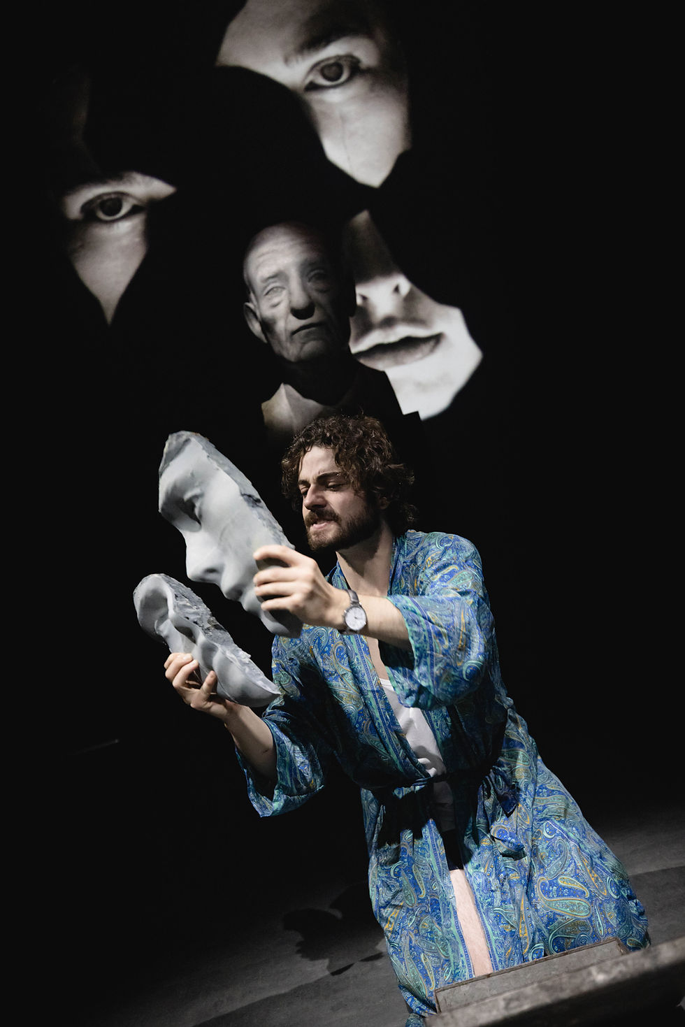 A man in a dressing gown holds pieces of a broke sculpture's face.
