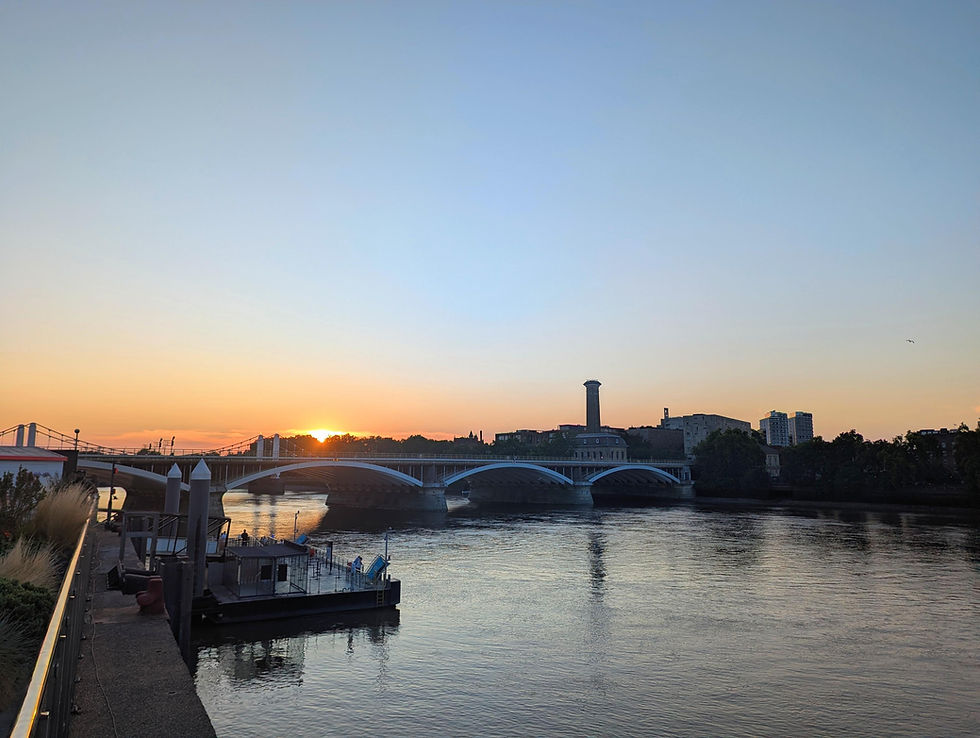 Sunset over a river with an arched bridge, city skyline, and calm water reflecting orange and blue sky.