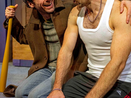 Two men kneel indoors, smiling and talking closely; one in a white tank top, the other holding a baseball bat, warm and intense.
