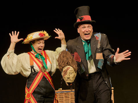 A woman throws her arms in the air as a man pulls a head from a basket