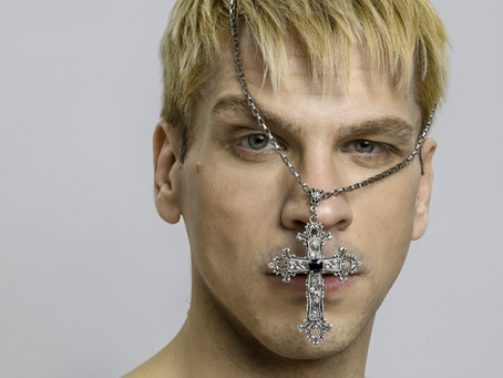 Close-up of a blonde man with a silver chain cross over his face, staring intensely against a plain grey background.