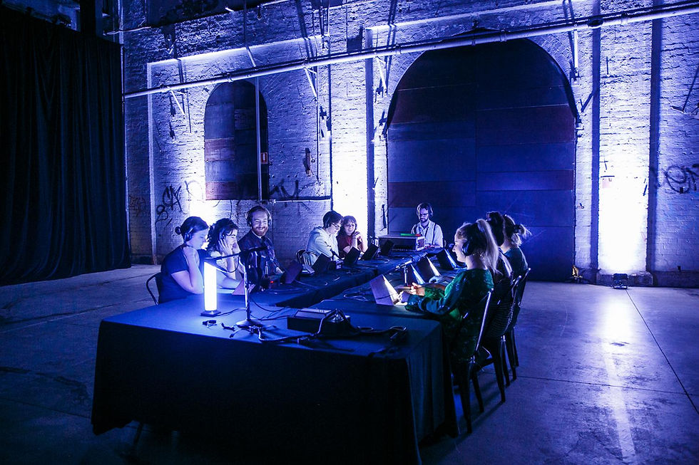 People wearing headphones work at laptops around long tables in a blue-lit industrial hall with brick arches.