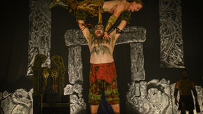 A strongman lifts a person above his head on a dimly lit stage with Norse-themed decor. Both wear warrior-like attire, exuding strength.