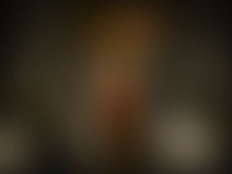 A strongman lifts a person above his head on a dimly lit stage with Norse-themed decor. Both wear warrior-like attire, exuding strength.