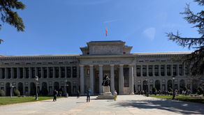 Front view of Museo del Prado under a clear blue sky. People walk and take photos near the statue in the courtyard. Trees frame the scene.
