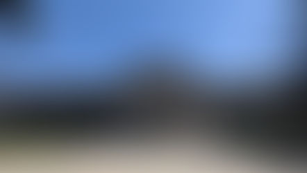 Front view of Museo del Prado under a clear blue sky. People walk and take photos near the statue in the courtyard. Trees frame the scene.